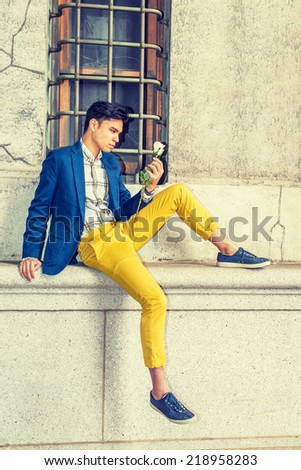 Man Missing You. Dressing in blue blazer, yellow pants, casual sneakers, a young handsome guy is sitting by a window on street, looking at white rose on hand, thinking.