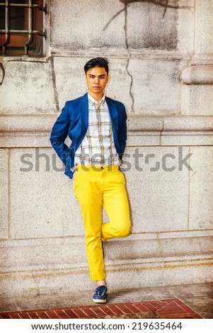 Man Relaxing Outside. Dressing in blue blazer, patterned under shirt, yellow pants, sneakers, hands on hips, a young guy is standing against old style wall, bending a leg, waiting, looking at you.