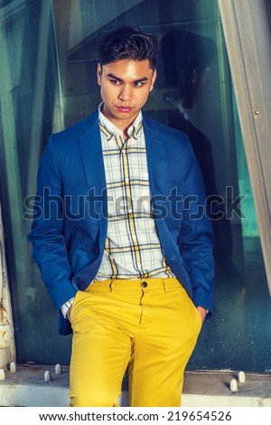 Man Thinking Outside. Dressing in blue blazer, pattern under wear, yellow pants, hands in pockets, a young guy is standing against glass wall on structure site, lowering head, sad, looking away.