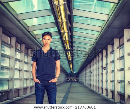 Wearing black V neck T shirt, blue jeans, wristwatch, two hands in pockets, a young guy standing inside indoor walkway with glass walls, ceiling, wooden floor, on campus.