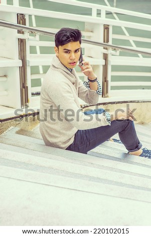 Man waiting for you. Dressing in kitted sweater with shawl collar cardigan, jeans, rolling over cuff, a young guy is sitting on stairs by fence, talking on mobile phone, looking back.