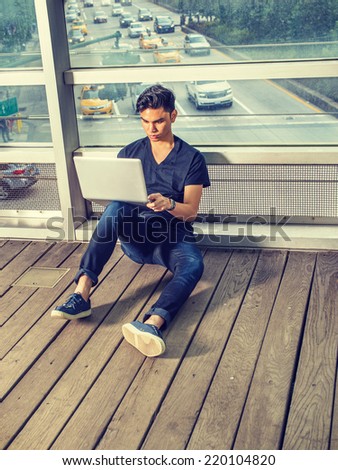 Busy Student. Wearing black V neck T shirt, blue jeans, sneakers, wristwatch, a young guy sitting on wooden floor, against glass wall, working on laptop computer. Cars on street in background.