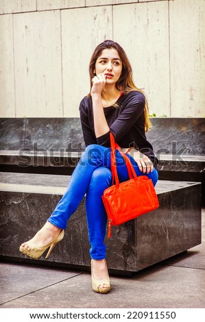Professional Woman Relaxing. Wearing black long sleeve sweater, blue pants, bracelets, arm carrying a red leather bag, a pretty lady is sitting on marble stone outside office building, relaxing.