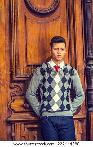 College Student. Wearing black, white, gray patterned sweater, red tie, blue jeans, hands on back, a young handsome guy is standing against an old fashion style office door, seriously looking at you.