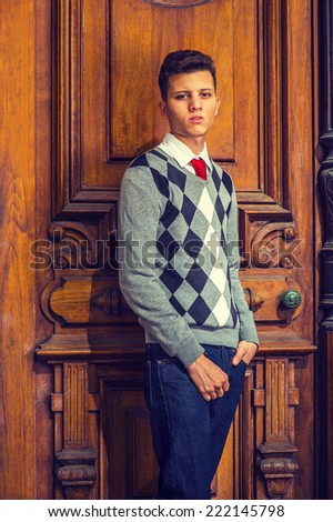 College Student. Wearing black, white, gray patterned sweater, red tie, blue jeans, a young handsome guy is standing by an old fashion style office door, seriously looking at you.