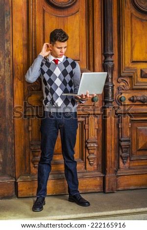 Man Working Outside. Wearing a patterned sweater, red tie, blue jeans, leather shoes, a young guy is standing outside office, looking down, scratching head, thinking, working on a laptop computer.