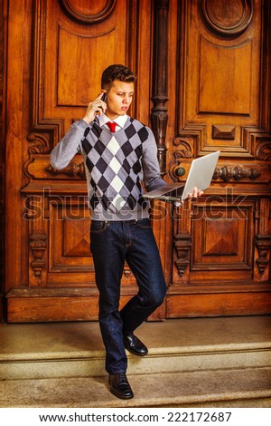 Man Working Hard. Wearing a patterned sweater, red tie, blue jeans, leather shoes, a young guy is waling down from office, talking on his mobile phone, working on a laptop computer in the same time.