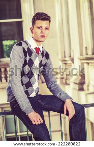 Portrait of College Student. Dressing in a black, white, gray patterned sweater, jeans, hands resting on laps, a young handsome businessman is sitting on a railing in office building, looking at you.