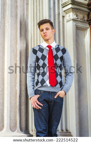 Portrait of College Student. Dressing in a black, white, gray patterned sweater, red tie, jeans, a young handsome businessman is standing by a pillar outside an office, looking at you.