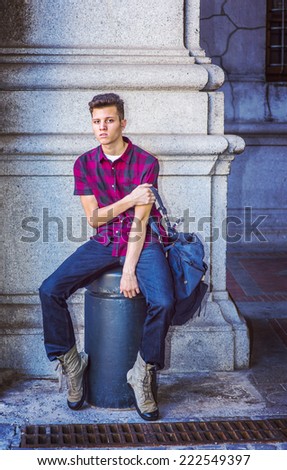 College Student on Street. Dressing in a short sleeve, black, red patterned shirt, jeans, boot shoes, carrying a shoulder bag, a young handsome guy is sitting on metal pillar on street, relaxing,
