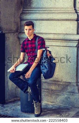 College Student. Dressing in short sleeve, black, red patterned shirt, jeans, boot shoes, carrying a shoulder bag, holding laptop computer, a young guy is sitting on metal pillar on street, relaxing,