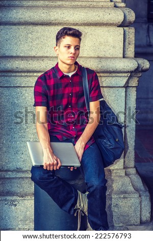 College Student. Dressing in short sleeve, black, red patterned shirt, jeans, carrying shoulder bag, holding laptop computer, a young guy is sitting on metal pillar on street, sad, relaxing, thinking.
