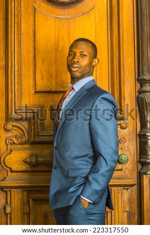 Portrait of Businessman. Dressing formally in blue suit, patterned shirt and necktie, wearing ear stud, short haircut, a young guy is standing in the front of an old style office door, looking at you.