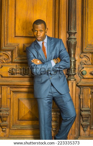 Dressing formally in blue suit, patterned undershirt, tie, short haircut, wooden bracelets, crossing arms, a young guy is standing in the front of an old style office door, confidently looking at you.