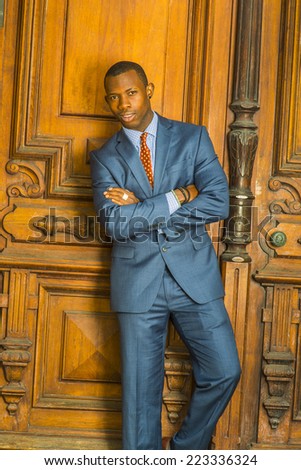 Dressing formally in blue suit, patterned undershirt, tie, short haircut, wooden bracelets, crossing arms, a young guy standing in the front of an old style office door, confidently looking at you.