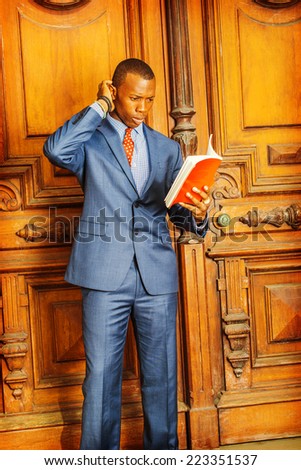 Man Thinking Hard. Dressing formally in blue suit, patterned undershirt, tie, short haircut, hand scratching his head, a young black professional is standing in an office, reading red book, thinking.