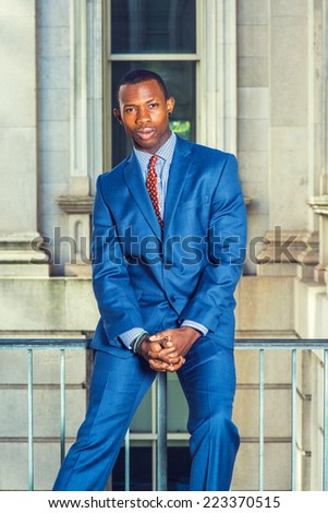 Portrait of Black Businessman. Dressing formally in blue suit, patterned undershirt, tie, short haircut, holding hands, a young handsome black guy is sitting on railing in office building, relaxing,