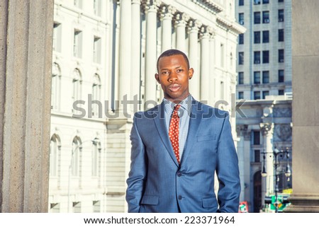 Portrait of Black Businessman. Dressing formally in blue suit, patterned undershirt, tie, short haircut, a young handsome black guy is standing in a business district, looking at you.