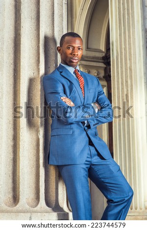 Business Man. Dressing formally in blue suit, patterned undershirt, tie, short haircut, crossing arms, a young black guy is standing by a column outside an office, relaxing, looking at you.