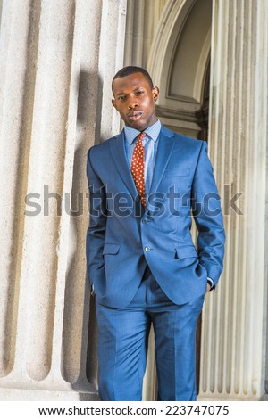 Business Man. Dressing formally in blue suit, patterned undershirt, tie, short haircut, hands in pockets, a young black guy is standing by a column outside an office, relaxing, waiting for you.