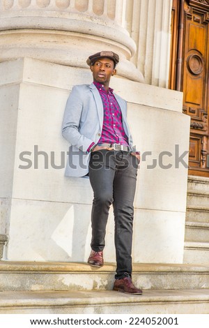 Man Urban Fashion. Wearing a Newsboy cap, dressing in light gray blazer, patterned pink, black under shirt, black pants, brown leather shoes, a young guy is leaning back on a column, relaxing.