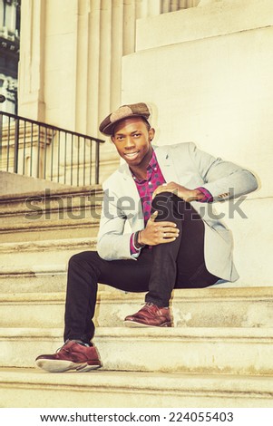 Man Urban Fashion. Wearing a Newsboy cap, dressing in light gray blazer, patterned pink, black under shirt, black pants, brown leather shoes, a young guy is sitting on stairs, smiling, relaxing.