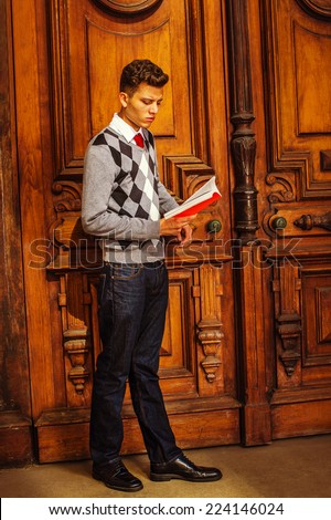 College Student. Wearing black, white, gray patterned sweater, red tie, blue jeans, leather shoes, a young handsome guy is standing by old fashion style office door, looking down, reading a red book.