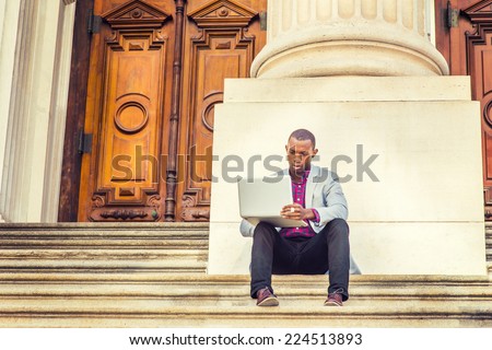 Dressing in light gray blazer, black pants, brown leather shoes, a young black guy is sitting on stairs, outside old fashion style office building, looking down, reading, working on laptop computer.