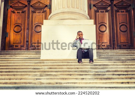 A young black business man is sitting on stairs of old fashion style office building door way, working on laptop computer and talking on his mobile phone in the same time. The Way to Success.