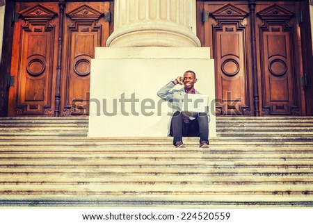 A young black business man is sitting on stairs of old fashion style office building door way, smiling, working on laptop computer, talking on his mobile phone. The Way to Success.