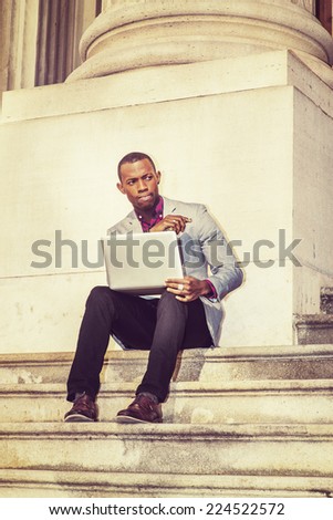 Man Working Outside. Dressing in light gray blazer, black pants, brown leather shoes,  short haircut, a young black guy is sitting on stairs outside office building, working on laptop computer.