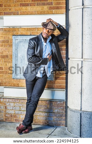 Man Urban Fashion. Wearing fashionable Newsboy cap, dressing in black blazer, pants, brown leather shoes, a young black guy is standing on the corner of the street, thinking, relaxing.