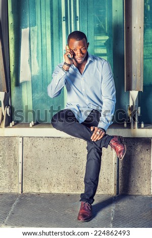 Man Calling Outside. Wearing a long sleeve shirt, black pants, brown leather shoes, short haircut, a young black guy is sitting on metal structures, crossing legs, talking on his mobile phone.