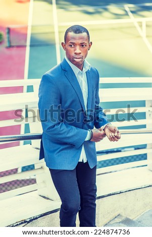Man Waiting for You. Wearing a blue blazer, black pants,, short haircut, a young black guy is standing against metal fences by a tennis court, hands touching cuff, looking away, lost in thought.