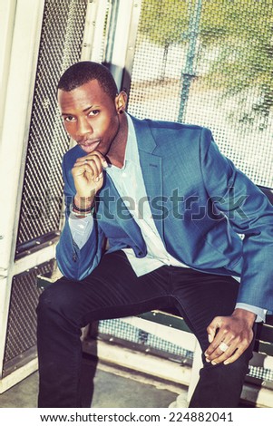 Wearing a blue blazer, black pants,, short haircut, ear stud,  bracelets, finger ring, a young black business man is sitting on bench outside, a hand touching his chin, bending over, thinking.