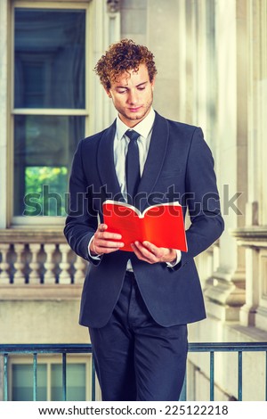 Man Reading Outside. Dressing in black suit with Shawl Lapel, black necktie, a young sexy guy with curly hair is standing inside office building, hands holding a red book, looking down, reading.