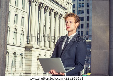 Businessman Working Outside. Dressing in black suit with Shawl Lapel, black necktie, a young guy with curly hair is standing in business district, hands holding computer, confidently looking forward.