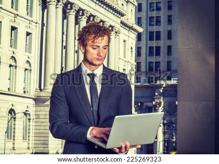 Businessman Working Outside. Dressing in black suit with Shawl Lapel, black necktie, a young sexy guy with curly hair is standing in business district, looking down, working on a computer.