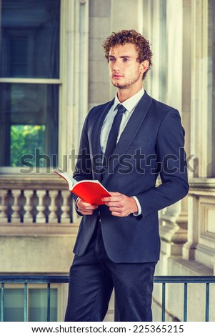 Man Reading Outside. Dressing in black suit with Shawl Lapel, black necktie, a young sexy guy with curly hair is standing inside office building, hands holding a red book, reading, looking forward.