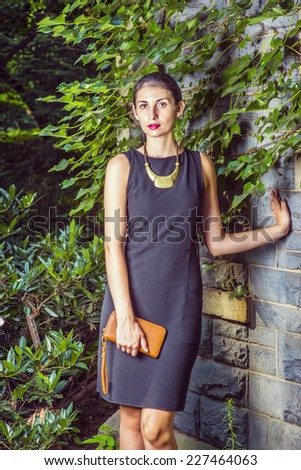 Working Woman Fashion. Dressing in black work dress, a necklace with golden pendant, holding small brown purse, a young sexy businesswoman is standing against a wall with ivy leaves, looking forward.