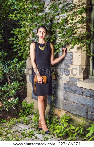 Working Woman Fashion. Dressing in black work dress, a necklace with golden pendant, holding small brown purse, a young sexy businesswoman is standing against a wall with ivy leaves, looking forward.