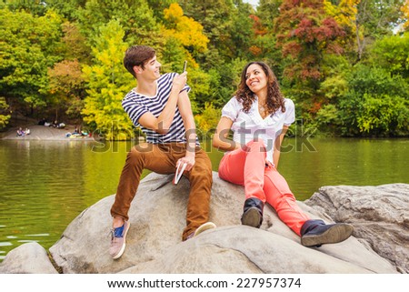 Young couple making fun by a lake in a park. Guy wearing a striped t shirt, brown pants, sneakers, holding a book, taking a picture. Girl dressing in white top, red pants, boot shoes, smiling.