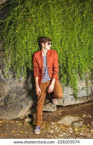 Man Casual Fashion. Wearing a dark reddish brown jacket, striped under shit, brown corduroy pants, sneakers, sunglasses, a young guy is standing against rocks wall with long leaves, waiting for you.