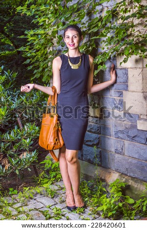 Working Woman Fashion. Dressing in black work dress, a necklace, a arm carrying a brown leather bag, a young sexy businesswoman is standing against a wall with ivy leaves, smiling, looking forward.