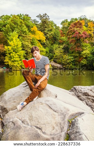 Man Reading Outside. Wearing striped T shirt, brown corduroy pants, sneakers, holding red book, a young handsome guy is sitting on rocks by a lake, looking down, reading. Autumn forest in background.