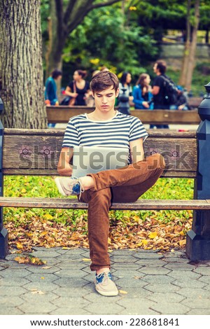 Man Working Outside. Wearing striped T shirt, brown corduroy pants, sneakers, a young handsome guy is sitting on bench on a park, crossing legs, looking down, reading on laptop computer.