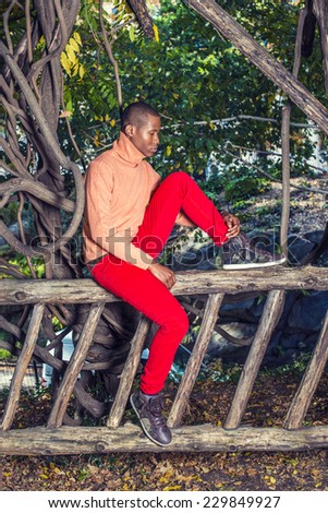 Thinking Outside. Dressing in light orange sweater with high collar, red pants, patterned boot shoes, a young black guy is sitting on fence, bending leg, looking down, thinking, lost in thought.