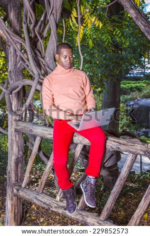 Man Working Outside. Dressing in light orange sweater with high collar, red pants, patterned boot shoes, a young black guy is sitting on wooden fence, looking down, reading on laptop computer.