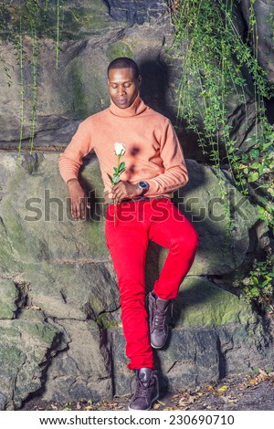 Man Missing You. Dressing in light orange sweater with high collar, red pants, boot shoes, wearing wristwatch, a young black guy is standing against rocks, holding white rose, looking down, thinking.
