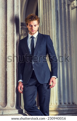 Man Thinking Outside. Dressing in black suit with Shawl Lapel, black necktie, a young sexy guy with curly hair is standing by column outside office, looking down, sad, thinking.